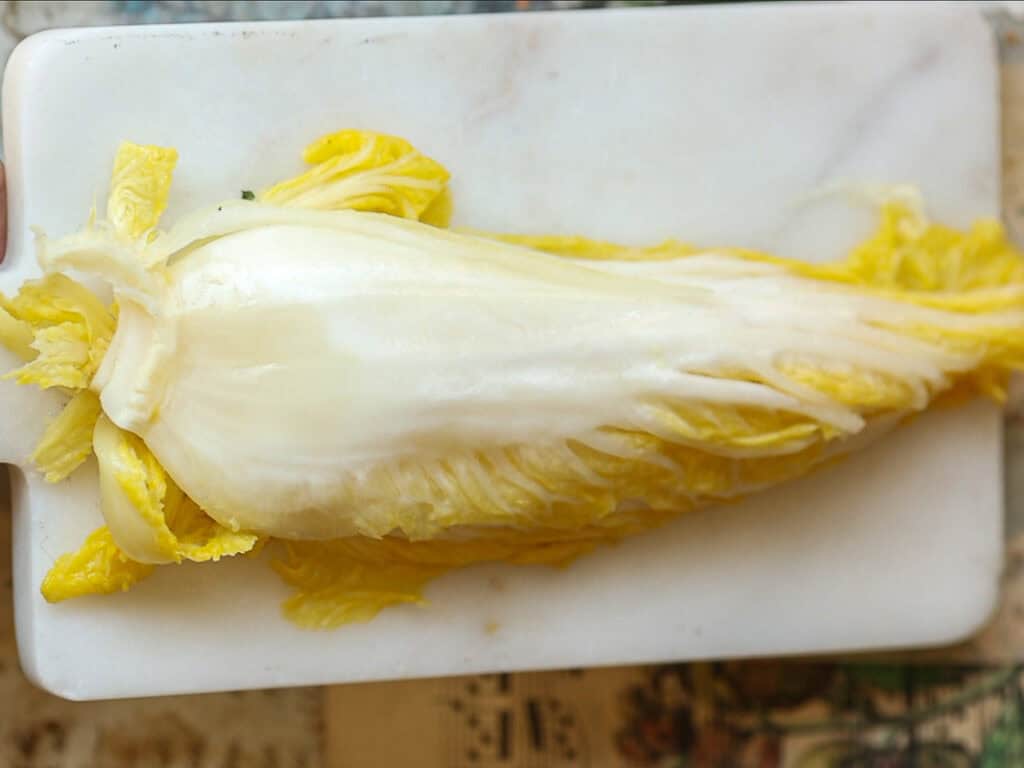 A half of napa cabbage with light yellow and white leaves lies on a white rectangular marble cutting board, showcasing its layers.